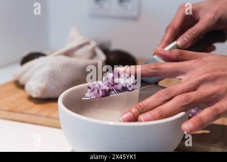 Par dessus la récolte anonyme femelle mettant l'oignon rouge haché dans un bol en céramique blanc tout en le coupant avec un couteau dans la cuisine pendant le processus de cuisson Banque D'Images