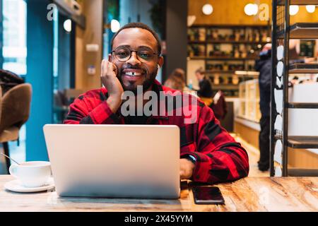 Un homme indépendant afro-américain positif qui navigue sur un netbook avec des écouteurs tout en travaillant à distance dans un café et en regardant un appareil photo Banque D'Images