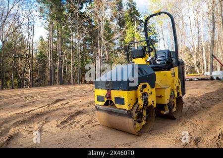 Gros plan du rouleau de compacteur jaune, un usiné utilisé pour le compactage du sol, de la surface d'asphalte, du gravier et du sable. Vue sur un bâtiment vide bordé d'arbres Banque D'Images