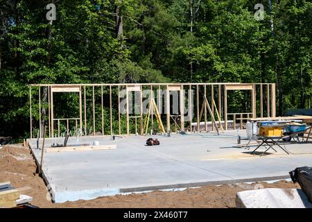 Nouveau chantier de construction de maison avec des montants en bois du premier mur en place, et des outils d'entrepreneurs sur la dalle de béton. Des arbres bordent l'arrière du bâtiment assis Banque D'Images