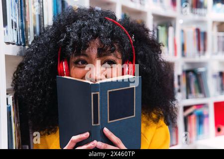 Jeune étudiante ethnique élégante et heureuse, avec de longs cheveux bouclés dans une tenue tendance et un casque au cou souriant tout en couvrant le visage avec un livre en M. Banque D'Images
