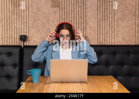 Jeune femme sérieuse dans des vêtements et des lunettes élégants et décontractés assis à la table avec un ordinateur portable et une boisson chaude et de mettre des écouteurs sans fil pendant la préparation Banque D'Images