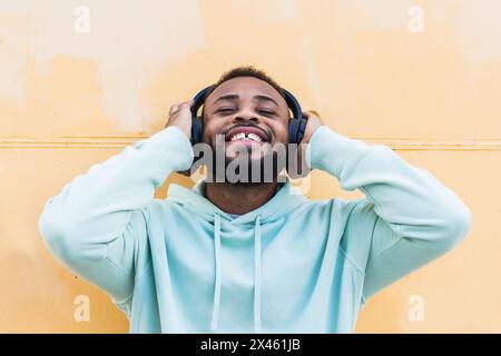 Joyeux adulte barbu afro-américain en bleu à capuchon appréciant musique avec casque sans fil sur fond jaune Banque D'Images