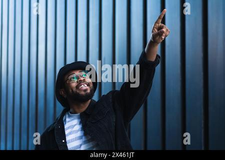 Souriant hippster afro-américain gars en chapeau et lunettes debout contre le mur d'un bâtiment moderne et pointant vers le haut Banque D'Images