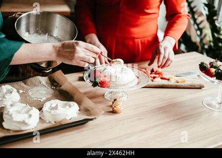 Un chef dans un tablier sarcelle façonne soigneusement des meringues sur une plaque de cuisson, entouré d'un cadre de cuisine animé avec des fruits frais comme l'ananas et l'orang Banque D'Images