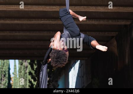 Une femme afro-américaine qui se retrouve à l'envers sur des lanières en deux parties tout en pratiquant le yoga aérien en ville Banque D'Images