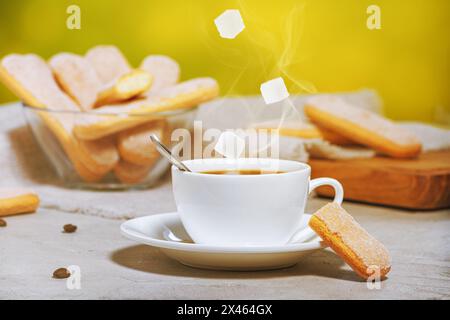 Tasse blanche de café aromatique chaud avec des cubes de sucre tombant et des biscuits traditionnels italiens savoiardi ou des biscuits ladyfinger sur une assiette dans un bol en verre Banque D'Images