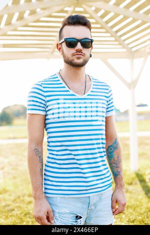 Trendy Handsome Hipster homme avec Une barbe dans des lunettes de soleil dans Une chemise à la mode se tient sur la plage. Banque D'Images