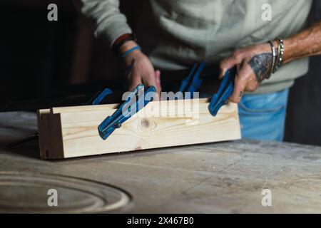 Croiser un menuisier mâle d'âge moyen méconnaissable avec des tatouages sur les mains dans les vêtements décontractés fixer des planches en bois avec métal réglable f colliers en atelier Banque D'Images