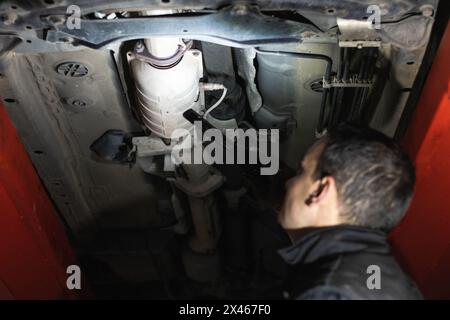 Vue latérale d'un mécanicien mâle méconnaissable qui vérifie le système d'échappement de la voiture relevée sur l'ascenseur dans le centre de réparation Banque D'Images