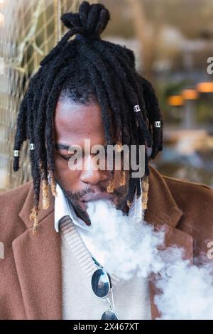 Boxer moderne pour adultes afro-américain avec une tenue tendance dreadlocks fume une cigarette électronique dans la rue Banque D'Images