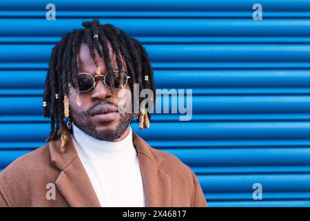 Cool Afro-américain mâle dans des lunettes de soleil élégantes et avec des dreadlocks debout contre le mur bleu de la ville Banque D'Images