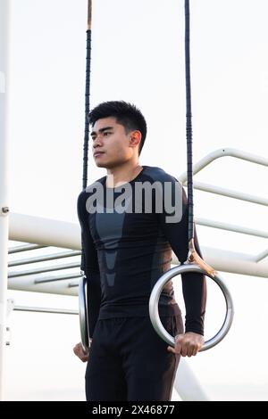 Petit angle de jeune sportif asiatique en casque d'activité noir sur les anneaux de gymnastique tout en effectuant des exercices sur le terrain de sport Banque D'Images