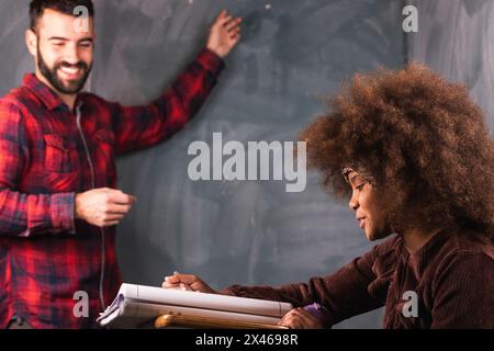 Professeur écrivant sur le tableau noir pour fille noire assise au bureau en classe pendant la leçon à l'école Banque D'Images