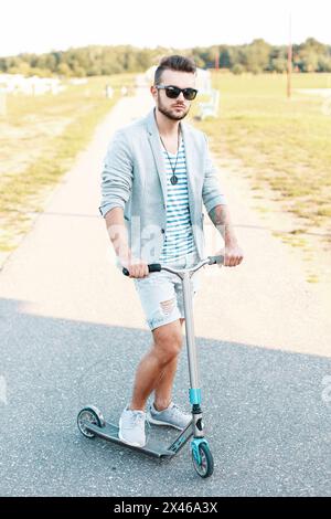 Élégant bel homme dans des vêtements à la mode chevauchant Un Kick Scooter sur la plage Banque D'Images