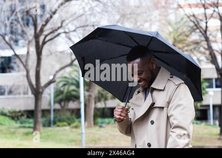 Homme afro-américain tendance adulte en trench élégant avec parapluie noir souriant et regardant l'appareil photo en position debout sur la rue urbaine dans Banque D'Images