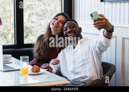 Couple multiracial heureux prenant un selfie tout en utilisant un ordinateur portable ensemble, assis à une table dans une pièce élégante à la maison Banque D'Images