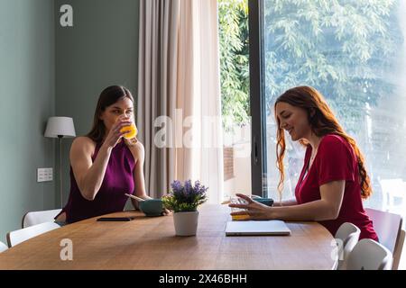 Jeunes amies joyeuses en tenues décontractées pour un délicieux déjeuner ensemble et bavardez tout en vous rassemblant à la table à manger dans la lumière appartement Banque D'Images