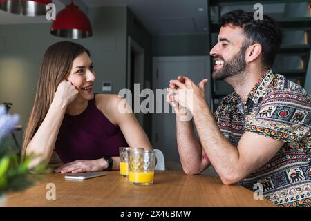 Jeune couple positif en vêtements décontractés ayant la conversation et appréciant jus d'orange frais tout en étant assis à la table à manger dans la cuisine Banque D'Images
