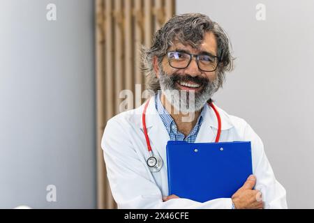 Joyeux homme âgé medic en uniforme et avec dossier debout dans la salle médicale dans la clinique et regardant la caméra Banque D'Images