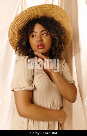 Une belle jeune femme afro-américaine aux cheveux bouclés pose gracieusement dans un studio, portant un chapeau de paille et une robe d'été. Banque D'Images