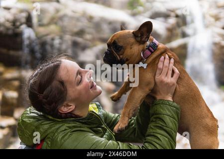 Vue latérale d'une rancheuse féminine enchantée qui embrasse un Bulldog français près d'une chute d'eau dans les bois tout en appréciant la randonnée dans les Pyrénées Banque D'Images