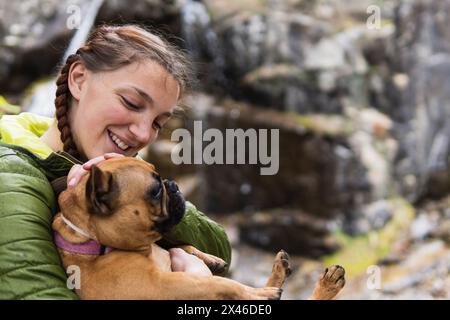 Vue latérale d'une rancheuse féminine enchantée qui embrasse un Bulldog français près d'une chute d'eau dans les bois tout en appréciant la randonnée dans les Pyrénées Banque D'Images