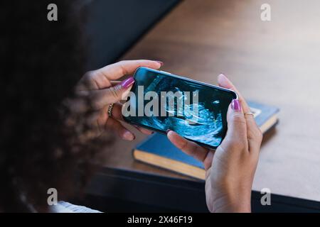 De dessus de la récolte femelle ethnique non reconnaissable assis à table avec une tasse de latte aromatique et livre et regardant bébé image d'ultrasons sur le téléphone cellulaire Banque D'Images