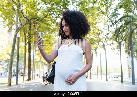 Femme afro-américaine positive avec une coiffure afro touchant le ventre tout en prenant le selfie sur le téléphone portable dans la rue avec des arbres verts Banque D'Images
