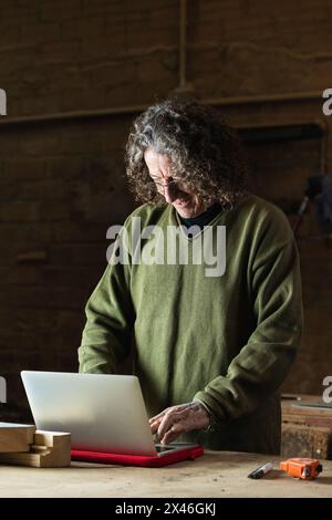 Artisan positif mature en lunettes dactylographiant sur ordinateur portable debout à l'établi en atelier avec des détails et des outils en bois Banque D'Images