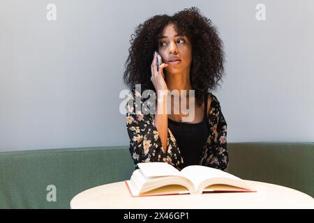 Jeune femme ethnique parlant au téléphone portable tout en étant assise sur un canapé et en regardant loin à table avec un manuel Banque D'Images