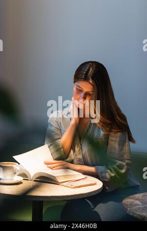 Jeune femme sereine se touchant le visage en lisant un manuel à la table de la cafétéria avec une tasse de boisson chaude Banque D'Images