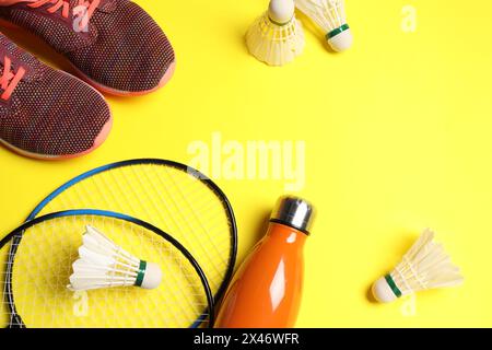 Volants de badminton en plume, raquettes, baskets et bouteille sur fond jaune, vue au-dessus. Espace pour le texte Banque D'Images