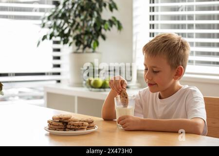 Garçon souriant alors qu'il trempe un biscuit fait maison dans un verre de lait, profitant de sa collation Banque D'Images