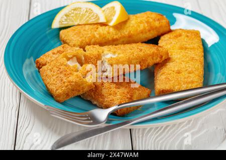 gros plan de filet de pollock d'alaska croustillant cuit au four sur un plat bleu avec des tranches de citron sur une table en bois blanc avec des couverts Banque D'Images