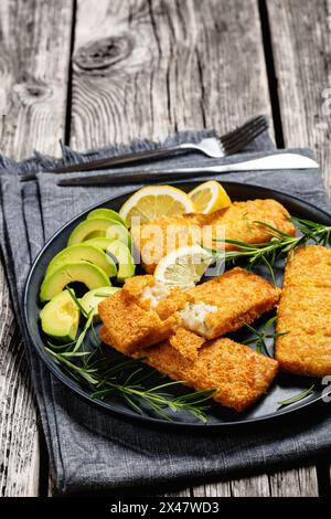 filet de pollock d'alaska pané croustillant sur plat noir avec des tranches de romarin, de citron et d'avocat sur table en bois rustique avec couverts, vue verticale Banque D'Images