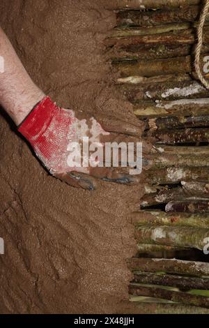 Personne avec la main gantée appliquant Daub à Un mur de ferraille dans la construction d'Une reconstruction d'une maison ronde de l'âge du fer, Royaume-Uni Banque D'Images