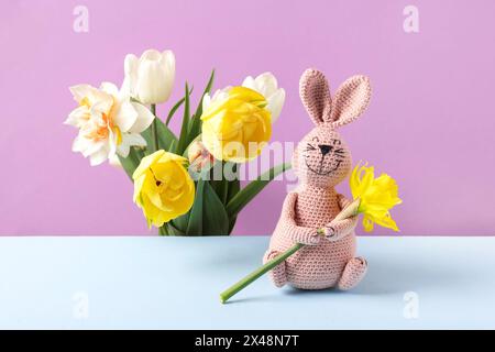 Lapin rose jouet tricoté avec jonquille jaune et bouquet de fleurs de printemps de tulipes et jonquilles sur table bleue et fond violet. Concept de spri Banque D'Images