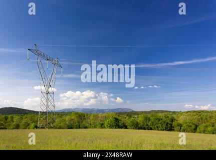 Champ vert contre un ciel bleu avec un pylône électricien au premier plan et des collines et des montagnes en arrière-plan Banque D'Images