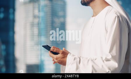 Authentique homme d'affaires musulman dans la traditionnelle Kandura blanche debout dans son bureau moderne, en utilisant smartphone à côté de la fenêtre avec des gratte-ciel. Concept d'homme d'affaires saoudien, émirati et arabe réussi. Banque D'Images