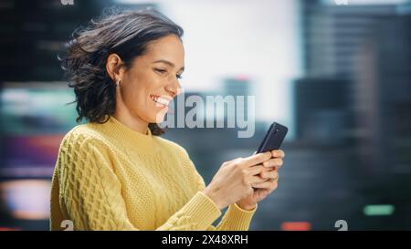 Street shot : Portrait de belle femme latine à l'aide d'un smartphone. Souriant hispanique entrepreneur féminin utilisant le téléphone portable pour le shopping en ligne, e-commerce heureux. Arrière-plan de mouvement flou. Banque D'Images