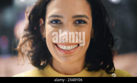 Gros plan Portrait de magnifique femme hispanique aux cheveux foncés avec des yeux bruns profonds regardant la caméra, souriant charmante. Heureuse jeune femme latine aime la vie avec plaisir, succès. Historique Bokeh City Street Banque D'Images