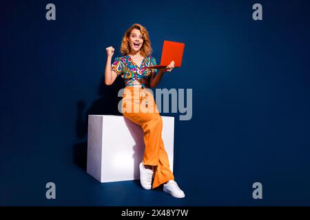 Photo pleine taille de jolie jeune femme tenir un ordinateur portable soulever le haut de poing d'usure isolé sur fond de couleur bleu foncé Banque D'Images