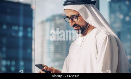 Homme d'affaires musulman prospère dans la tenue blanche traditionnelle debout dans son bureau moderne, en utilisant smartphone à côté de la fenêtre avec des gratte-ciel. Concept d'homme d'affaires saoudien, émirati et arabe réussi. Banque D'Images