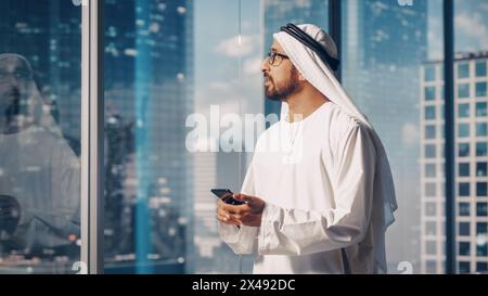 Authentique homme d'affaires musulman dans la traditionnelle Kandura blanche debout dans son bureau moderne, en utilisant smartphone à côté de la fenêtre avec des gratte-ciel. Concept d'homme d'affaires saoudien, émirati et arabe réussi. Banque D'Images