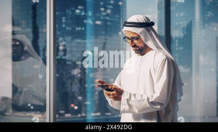 Homme d'affaires musulman prospère dans la tenue blanche traditionnelle debout dans son bureau moderne, en utilisant smartphone à côté de la fenêtre avec des gratte-ciel. Concept d'homme d'affaires saoudien, émirati et arabe réussi. Banque D'Images