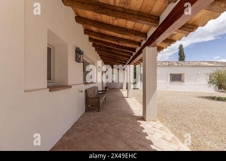 Porche couvert avec toit en bois d'une maison de campagne de style andalou avec des trottoirs en terre cuite, des bancs en bois, des murs blancs, un chemin de gravier, une nouvelle plante Banque D'Images