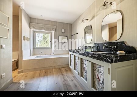 Une salle de bains spacieuse avec mobilier en bois avec un lavabo double, marbre noir, deux miroirs et une baignoire avec robinets de style vintage et windo en aluminium blanc Banque D'Images