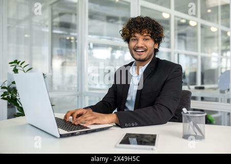 Homme d'affaires arabe gardant les mains sur le clavier de l'ordinateur portable tout en regardant la caméra avec le sourire dentelé. Employeur bouclé se sentant prêt à commencer une nouvelle semaine productive et rentable au bureau. Banque D'Images