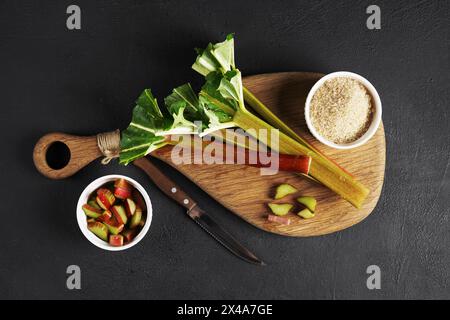 Rhubarbe hachée dans un bol, couteau sur une planche à découper en bois sur fond noir, vue de dessus. Banque D'Images